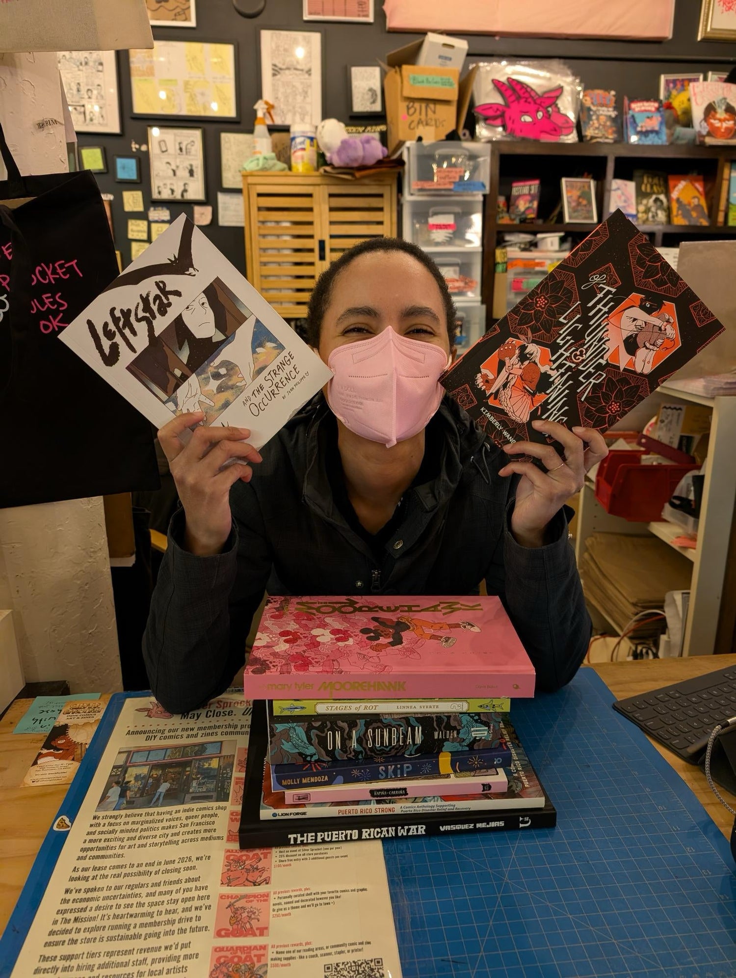Sol holds up some books next to their face and smiles under their mask over a pile of you guessed it more books.